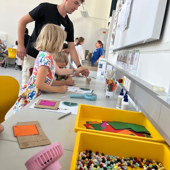 Ein Mann hilft zwei Kindern bei einem Bastelprojekt an einem Tisch mit bunten Papieren und Perlen. Hinter ihnen schaut eine Frau an eine Pinnwand.