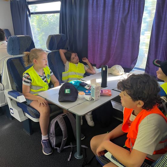 Eine Gruppe von Kindern in gelben Sicherheitswesten sitzt auf Sitzen in einem Zug, wobei ein Kind auf dem Sitz liegt und ein anderes zum Fenster schaut.