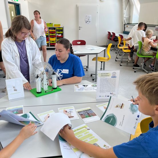 Eine Gruppe von Schülern und Lehrern in einem Klassenzimmer, ein Schüler benutzt ein wissenschaftliches Experiment, während andere Bücher und Materialien auf dem Tisch lesen.