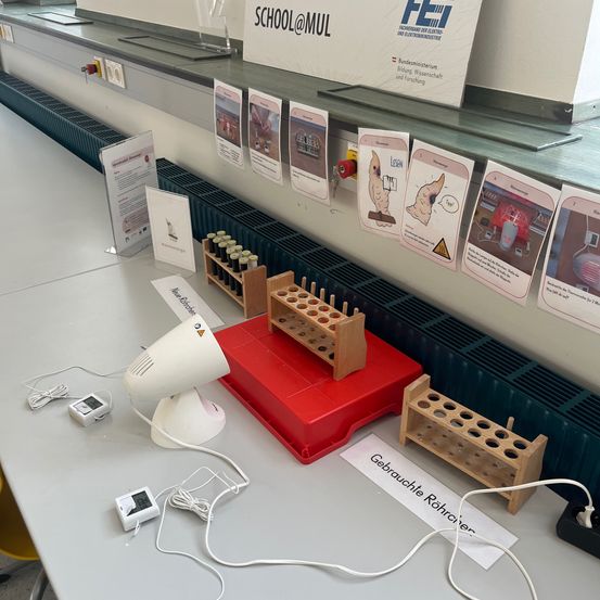 Ein wissenschaftliches Labor-Setup mit einer weißen Lampe, einem roten Tablett und verschiedenen Reagenzgläsern und Geräten. Ein Banner mit 'SCHOOL@MUL' und 'FEI' an der Wand.
