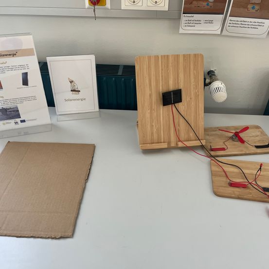 Ein holzernes Solarpanel treibt einen kleinen Ventilator auf einem weißen Tisch an. Kartonstücke liegen auf dem Tisch, und ein Heizkörper befindet sich dahinter. Papiere mit deutschem Text sind an der Wand befestigt.
