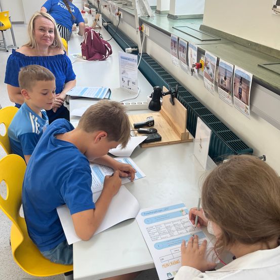 Drei Kinder in blauen Hemden sitzen an einem Tisch und schreiben auf Papier. Eine Frau sitzt hinter ihnen und beobachtet. Poster hängen an der Wand und ein Heizkörper ist daran montiert.