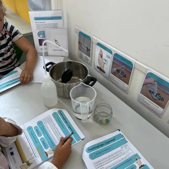 Zwei Kinder sitzen an einem Tisch mit einem Topf, einem Messbecher und einem Wasserglas. Ein Kind schreibt. Papiere mit Zeichnungen und Text sind auf dem Tisch. Es gibt Poster an der Wand.