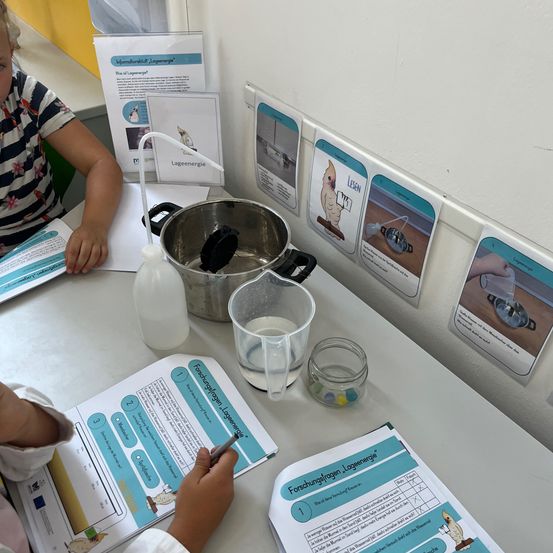Zwei Kinder sitzen an einem Tisch mit einem Topf, einem Messbecher und einem Wasserglas. Ein Kind schreibt. Papiere mit Zeichnungen und Text sind auf dem Tisch. Es gibt Poster an der Wand.