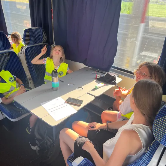 Eine Gruppe junger Personen in Warnwesten sitzt in einem Zug. Sie führen Gespräche, einige halten Papiere und ein Handy. Das Innere des Zuges hat blaue Sitze und Fenster.