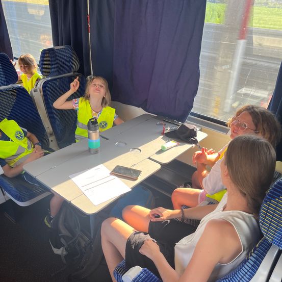Eine Gruppe junger Personen in Warnwesten sitzt in einem Zug. Sie führen Gespräche, einige halten Papiere und ein Handy. Das Innere des Zuges hat blaue Sitze und Fenster.