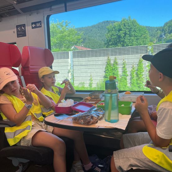 Drei Kinder in gelben Westen sitzen in einem Zug. Sie essen Snacks von einem Tablett auf dem Tisch. Eine Wasserflasche steht auf dem Tisch. Das Fenster des Zuges zeigt eine Aussicht auf einen Zaun, Bäume und Berge.