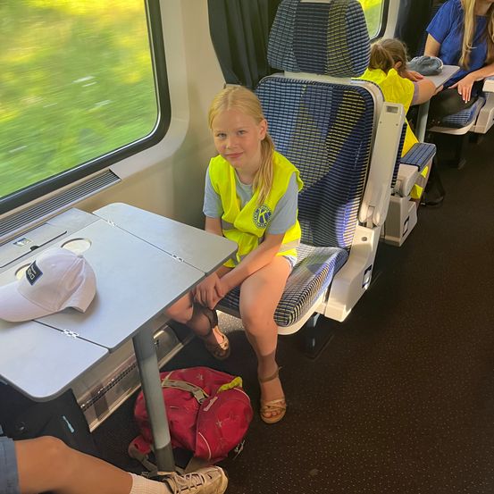 Ein Mädchen in einer gelben Sicherheitsweste sitzt lächelnd in einem Zug. Neben ihr ruht ein Kind. Hinter ihnen sitzt eine Frau. Vor dem Fenster verwischt sich vorbeiziehende grüne Landschaft.