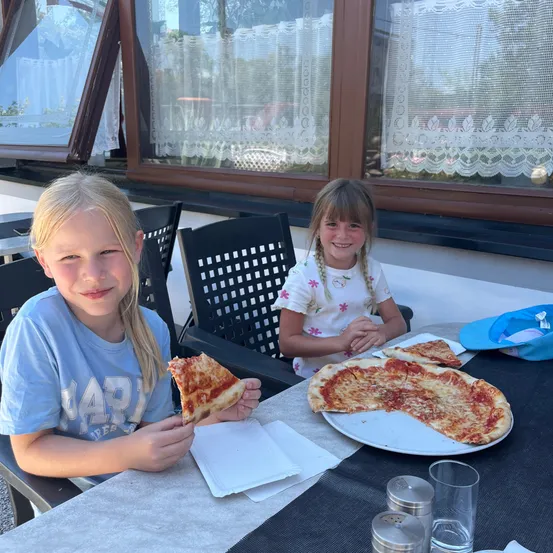 Zwei Mädchen essen Pizza an einem Tisch in einem Restaurant. Eines hält eine Scheibe, während das andere zusieht. Eine blaue Kappe und Teller mit Pizza sind auf dem Tisch.