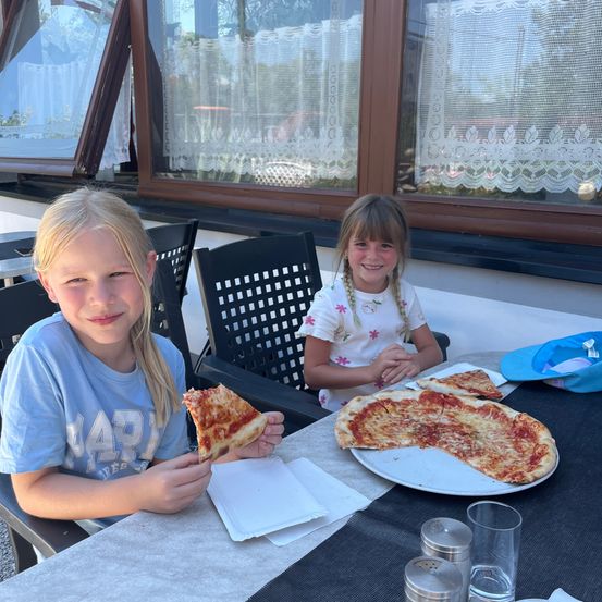 Zwei Mädchen essen Pizza an einem Tisch in einem Restaurant. Eines hält eine Scheibe, während das andere zusieht. Eine blaue Kappe und Teller mit Pizza sind auf dem Tisch.