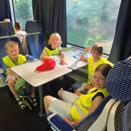 Vier Kinder sitzen in einem Zug und tragen reflektierende Westen. Sie scheinen fröhlich zu sein, mit einer roten Kappe auf dem Tisch vor ihnen. Die Fenster zeigen eine verschwommene Aussicht nach draußen.