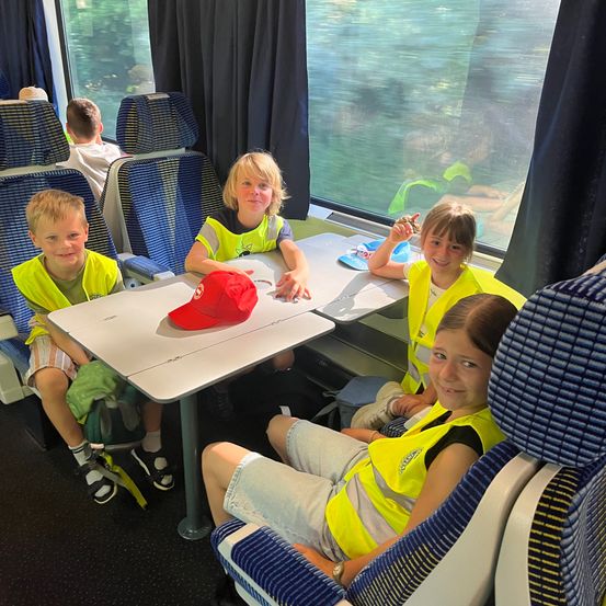 Vier Kinder sitzen in einem Zug und tragen reflektierende Westen. Sie scheinen fröhlich zu sein, mit einer roten Kappe auf dem Tisch vor ihnen. Die Fenster zeigen eine verschwommene Aussicht nach draußen.