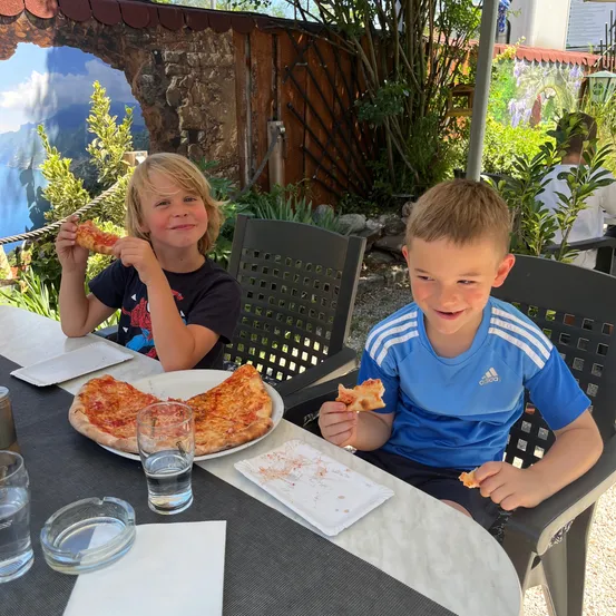 Zwei Jungen essen Pizza an einem Tisch. Einer hält eine Scheibe, während der andere eine Pizza hält. Auf dem Tisch stehen Teller, Gläser und eine Aschenschale.