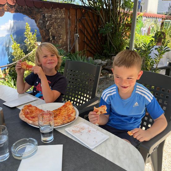 Zwei Jungen essen Pizza an einem Tisch. Einer hält eine Scheibe, während der andere eine Pizza hält. Auf dem Tisch stehen Teller, Gläser und eine Aschenschale.