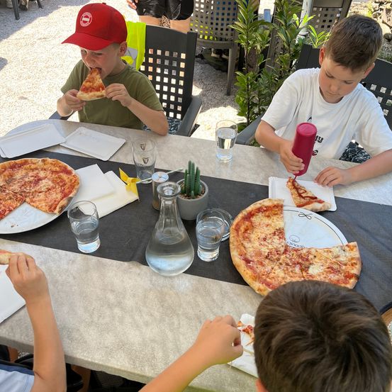 Vier Kinder essen Pizza an einem Tisch. Zwei Pizzen sind auf dem Tisch, während ein Kind eine Scheibe isst. Ein anderes Kind fügt einer Scheibe Käse hinzu.