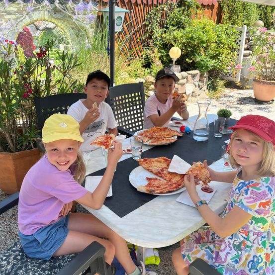 Vier Kinder sitzen um einen Tisch im Freien und essen Pizza. Zwei Mädchen und zwei Jungen. Sie lächeln und scheinen ihre Mahlzeit zu genießen.