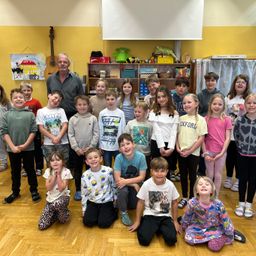 Eine Gruppe von Kindern und ein erwachsener Mann stehen in einem Klassenzimmer. Sie alle lächeln für ein Foto. Ein Holzregal mit Spielzeug und einer Gitarre ist im Hintergrund.