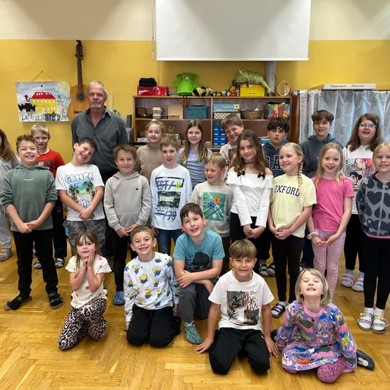 Eine Gruppe von Kindern und ein erwachsener Mann stehen in einem Klassenzimmer. Sie alle lächeln für ein Foto. Ein Holzregal mit Spielzeug und einer Gitarre ist im Hintergrund.