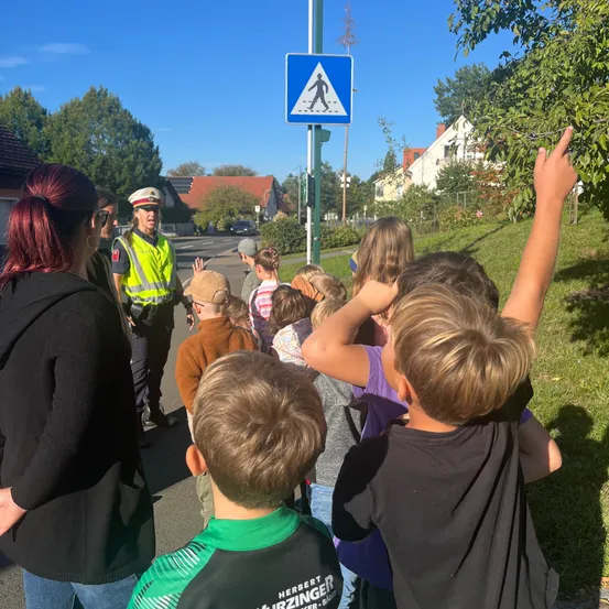 Eine Gruppe von Kindern und Erwachsenen steht am Straßenrand, wobei ein Verkehrspolizist in einer gelben Weste auf ein Fußgängerübergangsschild zeigt.