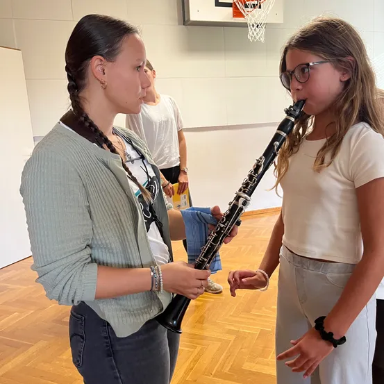 Eine Frau mit geflochtenen Haaren lehrt ein junges Mädchen, wie man Klarinette spielt, in einem Raum mit Holzböden und einem Basketballkorb im Hintergrund.