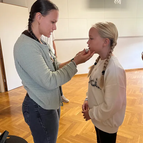 Eine Frau mit geflochtenen Haaren steht in einem Raum und scheint einem jungen Mädchen mit blonden Haaren Make-up aufzutragen.