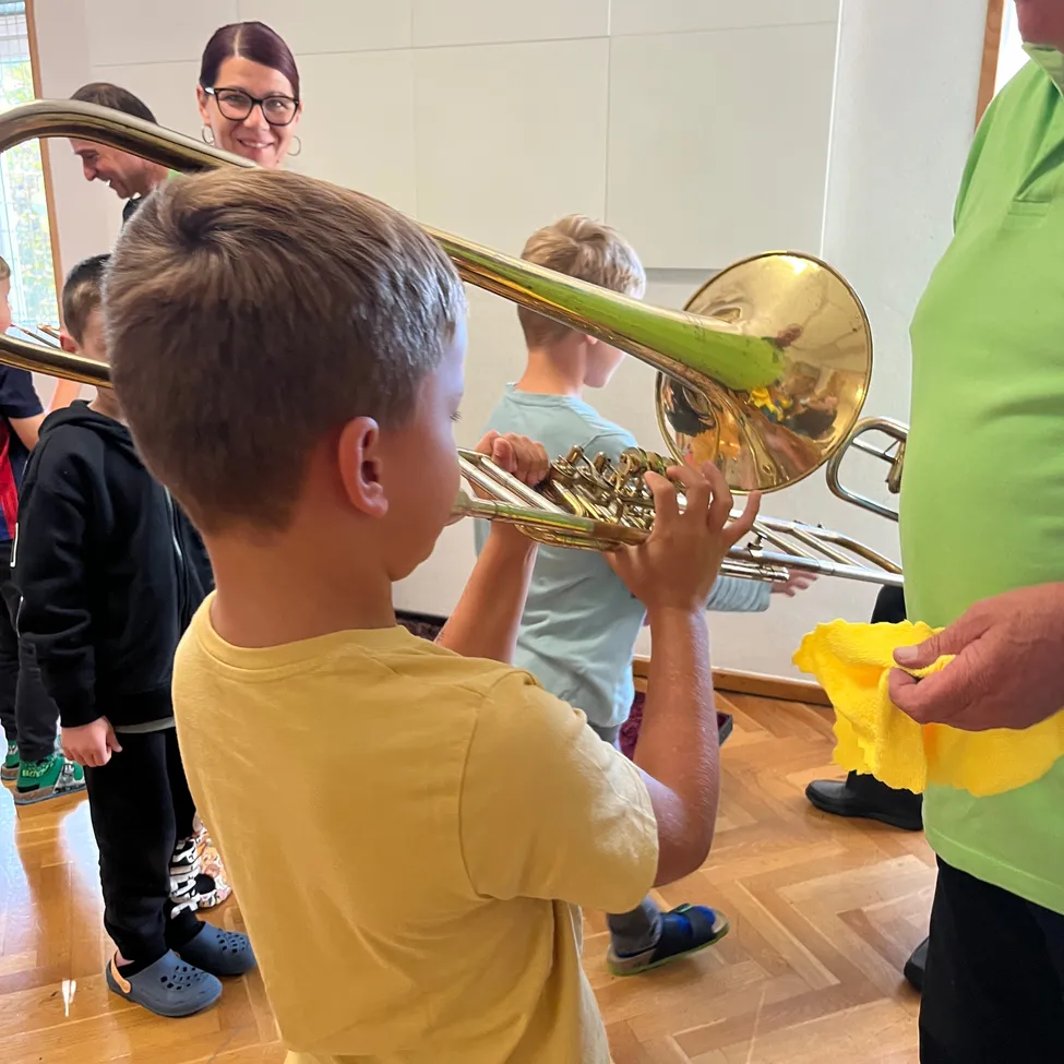 Ein junger Junge spielt ein Tenorhorn, während ein Mann ein gelbes Tuch hält und eine Frau zuschaut.