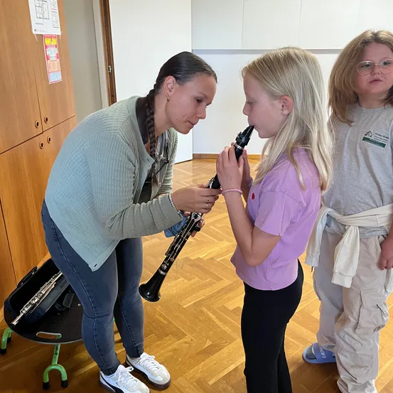 Eine Frau hilft einem jungen Mädchen, ein Klarinette zu spielen. Das Mädchen hält das Klarinette an den Mund, und die Frau hält den Hals des Klarinettes. Sie stehen auf einem Holzboden.