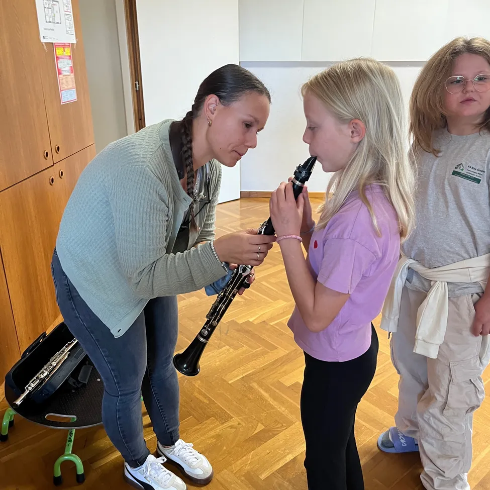 Eine Frau hilft einem jungen Mädchen, ein Klarinette zu spielen. Das Mädchen hält das Klarinette an den Mund, und die Frau hält den Hals des Klarinettes. Sie stehen auf einem Holzboden.