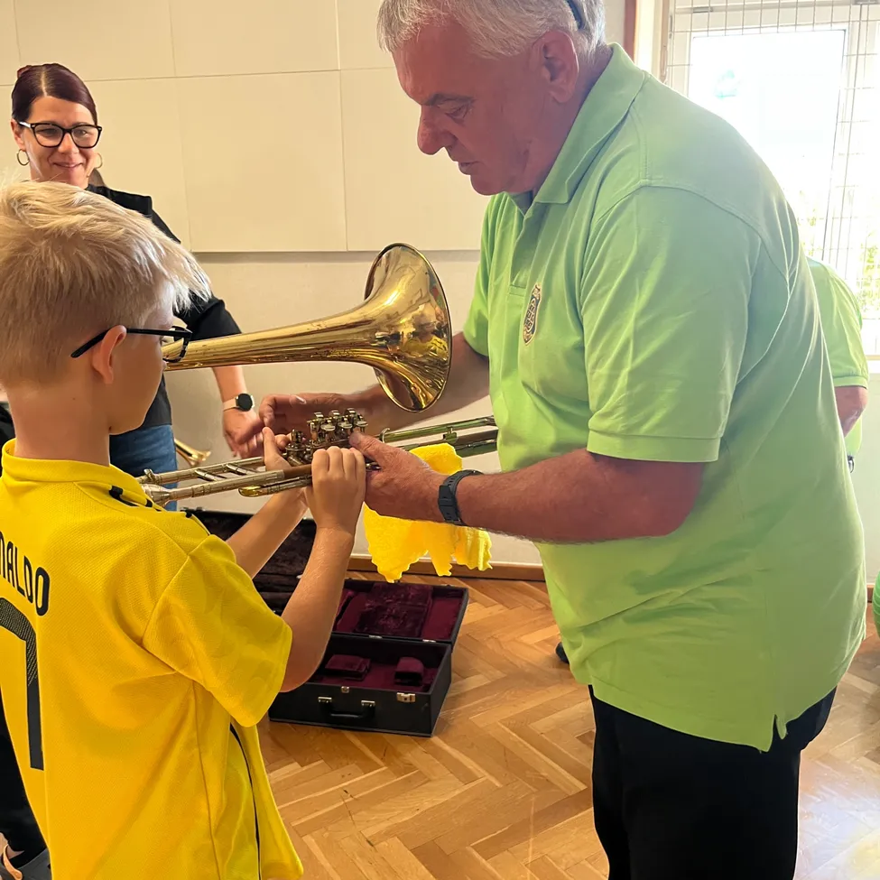 Ein älterer Mann in einem grünen Poloshirt hilft einem jungen Jungen in einem gelben Shirt, ein Tenorhorn zu spielen.