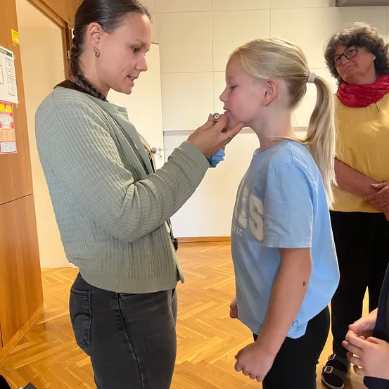 Eine Frau mit geflochtenen Haaren untersucht die Zähne eines jungen Mädchens mit einem Zahnspiegel. Das Mädchen trägt ein blaues Shirt mit weißen Buchstaben. Hinter ihnen steht eine Frau mit Brille und einem roten Schal.