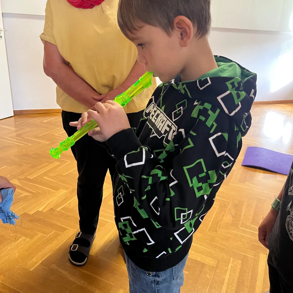 Ein junger Junge spielt eine grüne Blockflöte in einem Raum mit Holzboden. Eine Frau in einem gelben Shirt und einem roten Schal steht hinter ihm.