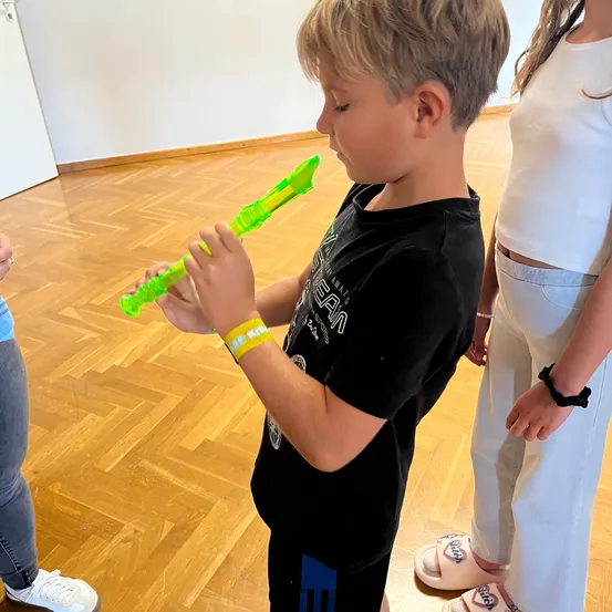 Ein kleiner Junge spielt eine grüne Flöte in einem Raum mit Holzboden. Er trägt ein schwarzes T-Shirt und Shorts. Hinter ihm steht ein Mädchen in weißen Hosen und Hausschuhen und schaut zu.