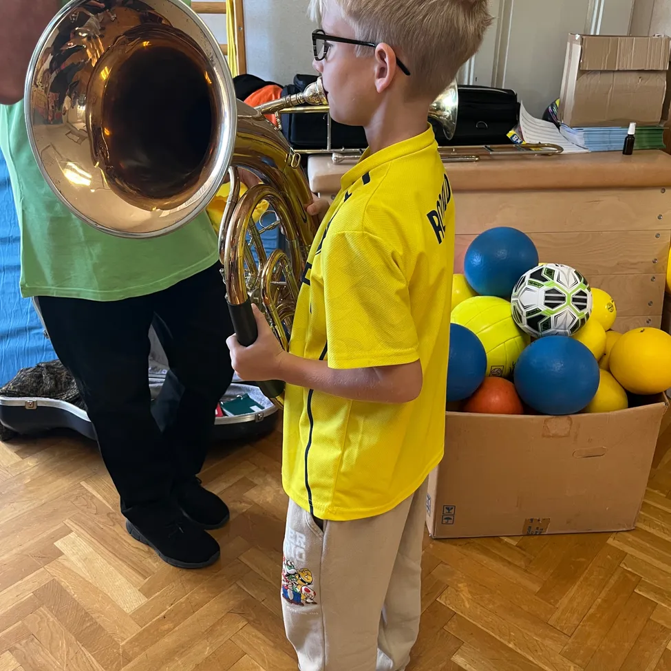 Ein junger Junge spielt eine Tuba, während ein Mann in einem grünen Shirt hinter ihm steht.