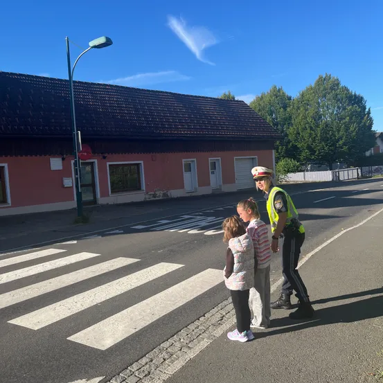 Eine Verkehrspolizistin steht am Straßenrand und spricht mit zwei jungen Mädchen an einem Zebrastreifen.