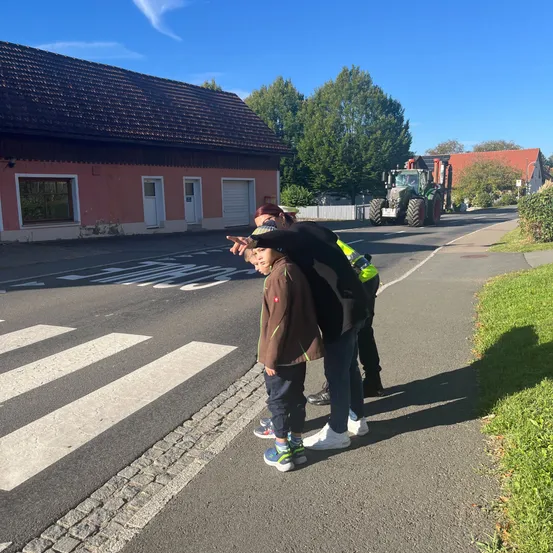 Ein Mann und ein Kind stehen auf einem Bürgersteig und zeigen auf einen Zebrastreifen. Dahinter fährt ein Traktor auf der Straße.