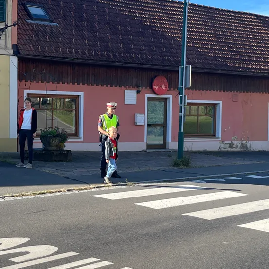 Eine Frau und ein Kind überqueren die Straße mit Hilfe eines Verkehrspolizisten. Die Szene spielt vor einem Gebäude mit einem roten Schild und einer Straßenlaterne.