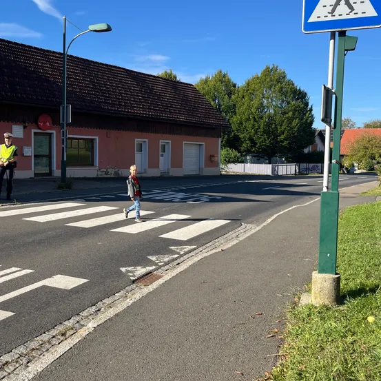 Ein Kind überquert eine Straße mit einem Zebrastreifen und einem Verkehrswächter im Hintergrund.