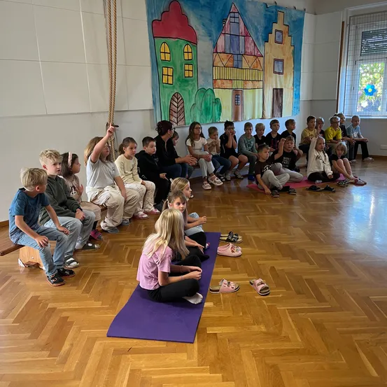 Eine Gruppe von Kindern sitzt auf dem Boden in einem Raum, einige tragen Schuhe, und eine Frau steht vor ihnen.