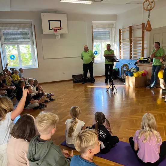 Eine Gruppe von Kindern sitzt auf dem Boden in einem Raum und hört sich eine Präsentation von zwei Männern und einer Frau an.