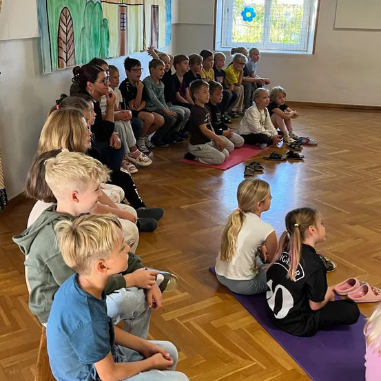 Eine Gruppe von Kindern und Erwachsenen sitzt auf dem Boden in einem Raum. Sie schauen auf etwas vor ihnen.