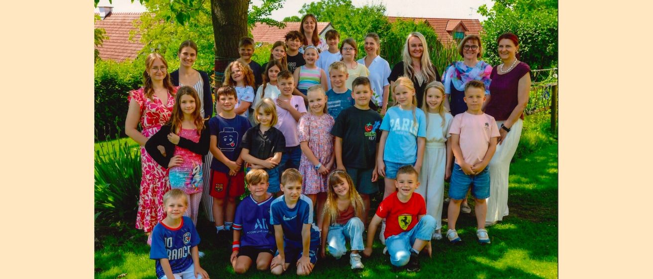 Ein Gruppenfoto von Kindern und Erwachsenen auf einem Rasenfeld, mit einem Baum und Häusern im Hintergrund.