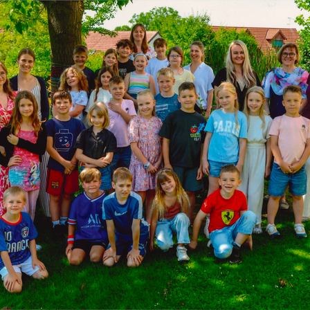 Eine große Gruppe von Kindern und Erwachsenen posiert für ein Foto im Garten, einige tragen Brillen und Turnschuhe. Sie stehen und hocken auf dem Rasen mit Bäumen und Häusern im Hintergrund.