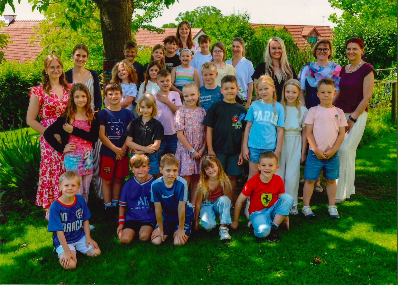 Eine große Gruppe von Kindern und Erwachsenen posiert für ein Foto im Garten, einige tragen Brillen und Turnschuhe. Sie stehen und hocken auf dem Rasen mit Bäumen und Häusern im Hintergrund.