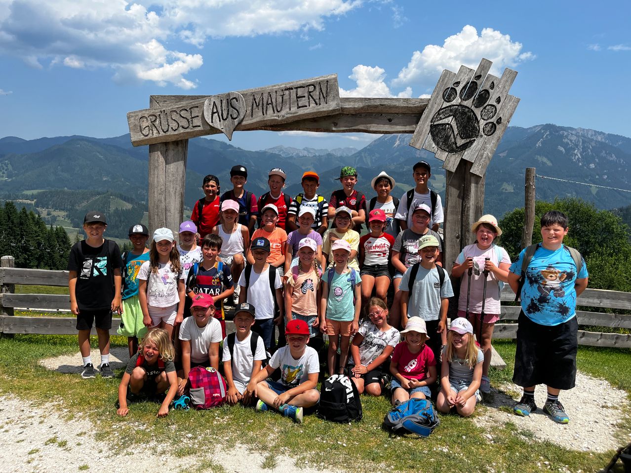 Eine Gruppe von Kindern vor einem Holzschild mit 'Grüsse aus Mauntern', das Foto vor einer malerischen Bergkulisse macht.