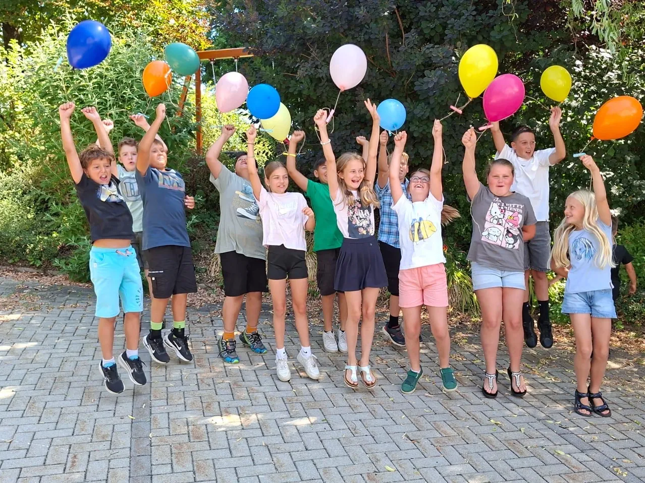 Eine Gruppe von Kindern, die mehrfarbige Luftballons halten, springen nach oben, wobei einige Ballons in der Luft schweben. Sie stehen auf einem Ziegelboden, mit Bäumen und Grünpflanzen hinter ihnen.