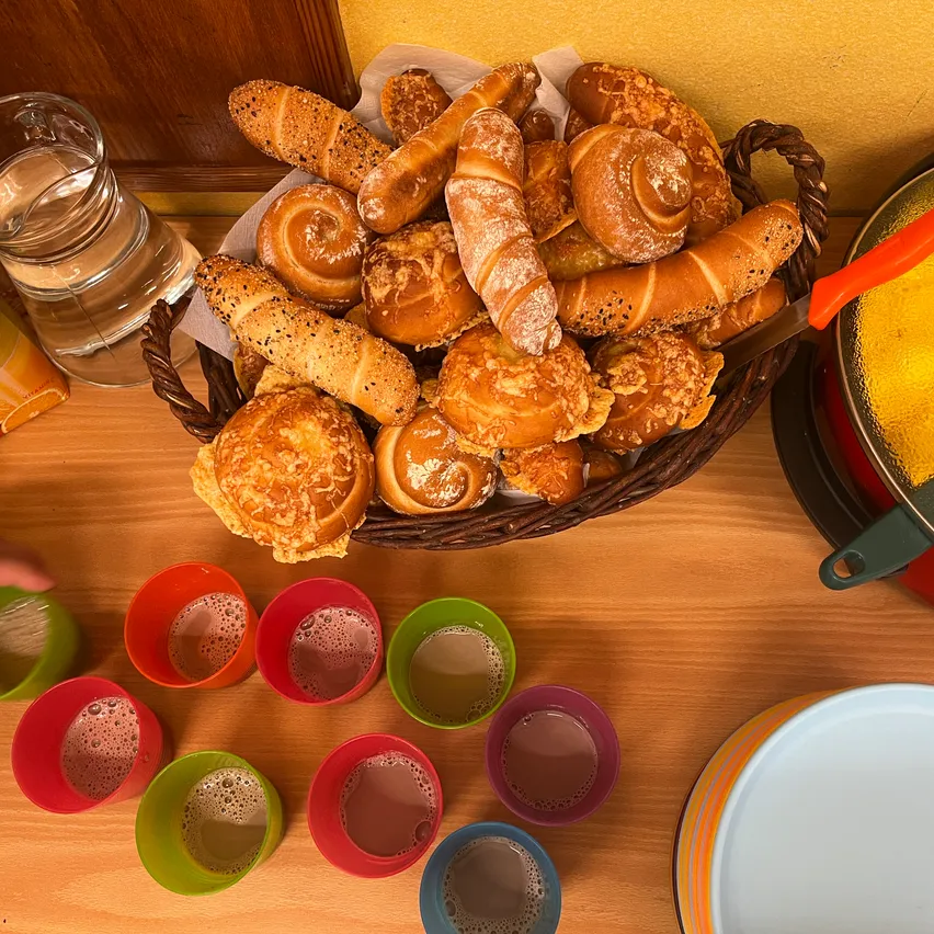 Ein Holztisch zeigt einen Korb, der mit verschiedenen Arten von Brot gefüllt ist. Das Brot hat verschiedene Formen und Größen, wobei einige Sesamkörner auf der Oberseite haben. Es gibt auch mehrere Tassen mit möglicherweise Kaffee. Ein Wasserkrug befindet sich auf der linken Seite.