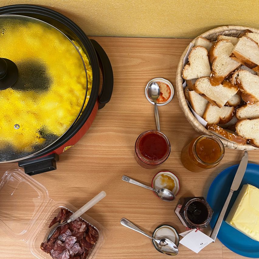 Ein Holztisch zeigt einen Topf mit Suppe, einen Korb mit Brot, einen Teller mit Butter, ein Messer und verschiedene Gläser mit Beilagen.