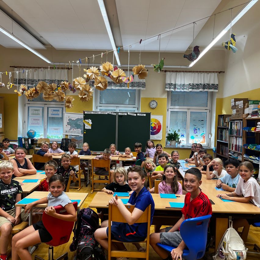 Ein Klassenzimmer voller Kinder, die an Schreibtischen sitzen. Der Raum ist hell mit natürlichem Licht und mit Papiervögeln dekoriert. Die Kinder lächeln und sind engagiert.