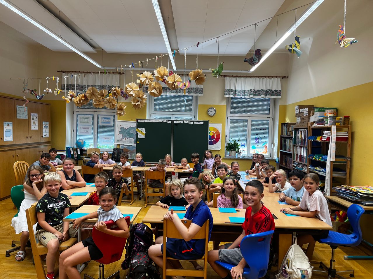 Ein Klassenzimmer voller Kinder, die an Schreibtischen sitzen. Der Raum ist hell mit natürlichem Licht und mit Papiervögeln dekoriert. Die Kinder lächeln und sind engagiert.