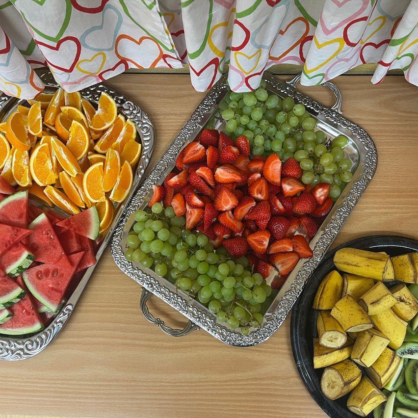 Auf einem Holztisch stehen zwei Tabletts mit Früchten: eines mit geschnittenen Orangen und Wassermelonen, das andere mit Erdbeeren und grünen Trauben. Ein weiteres Teller hält Bananen und Kiwis.